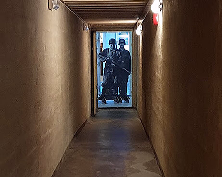 En smal, mørk korridor med grove vegger og betonggulv, som leder til en døråpning med blått lys og figurer av soldater i uniform.