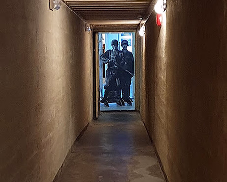 Lang, smal betongkorridor som leder mot to figurer i militæruniformer i en døråpning