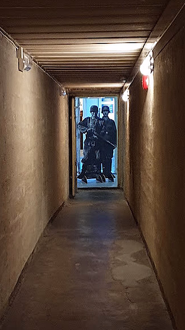 En lang, smal og dunkel gang med grove vegger og tretak. I enden er en døråpning opplyst i blått lys, med figurer av soldater i uniform.