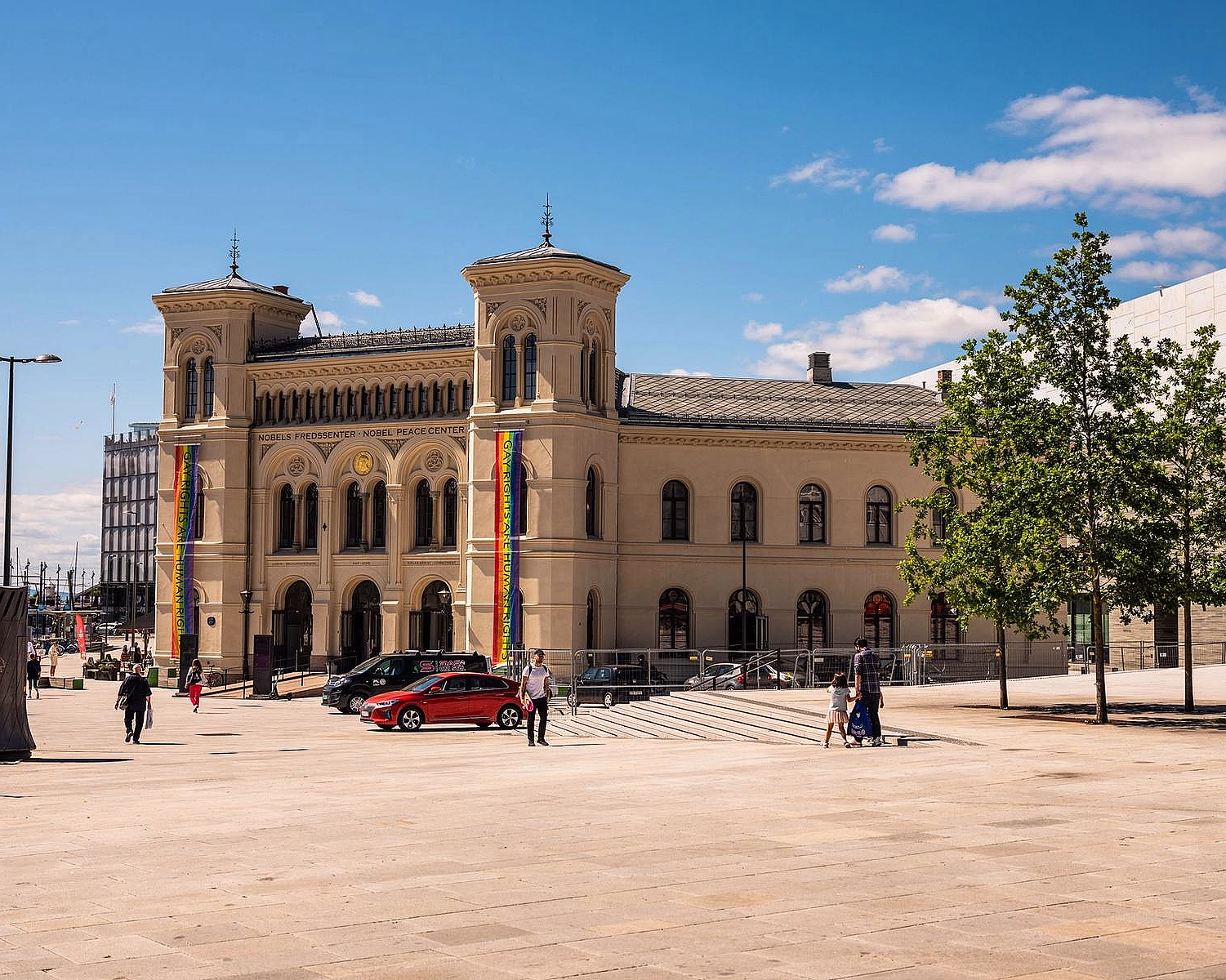 Nobels Fredssenter, en beige steinbygning med to tårn og regnbuebannere, sett fra en stor plass under en blå himmel.