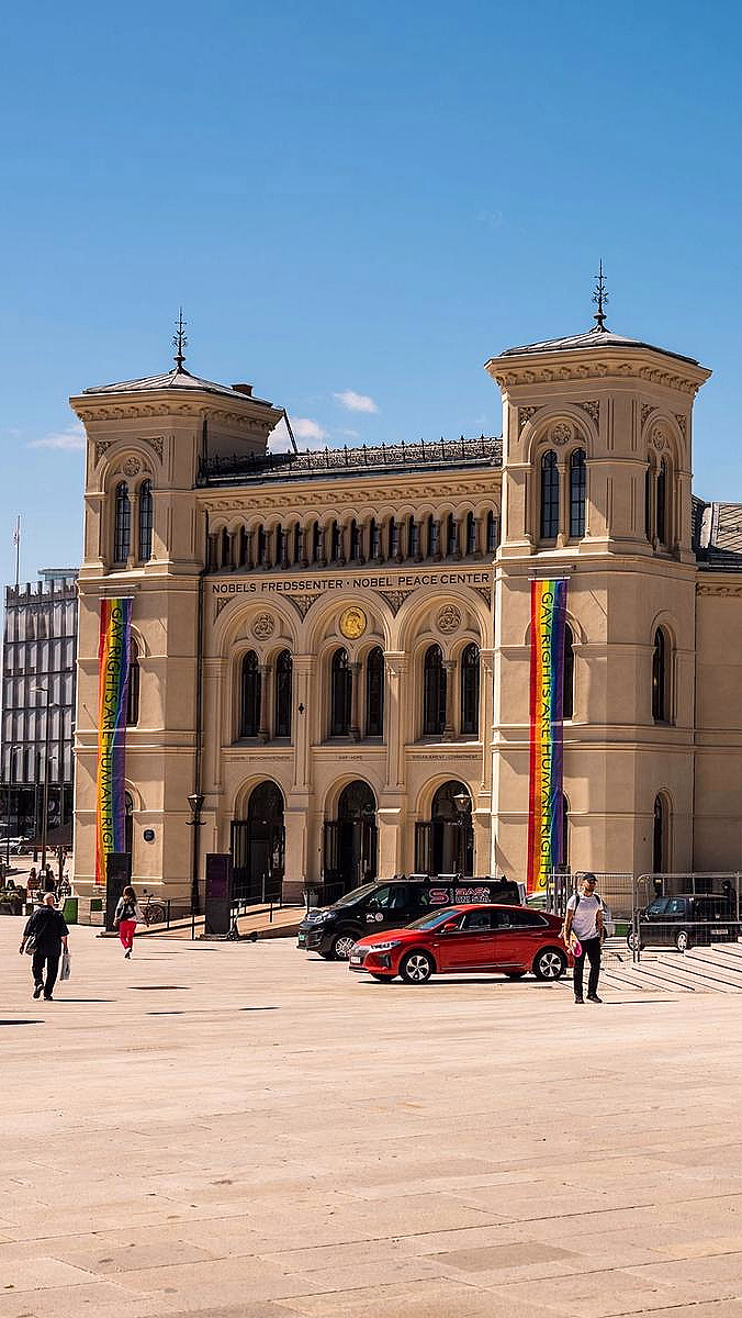 Nobels Fredssenter, en beige steinbygning med to tårn og regnbuebannere, sett fra en stor plass under en blå himmel.