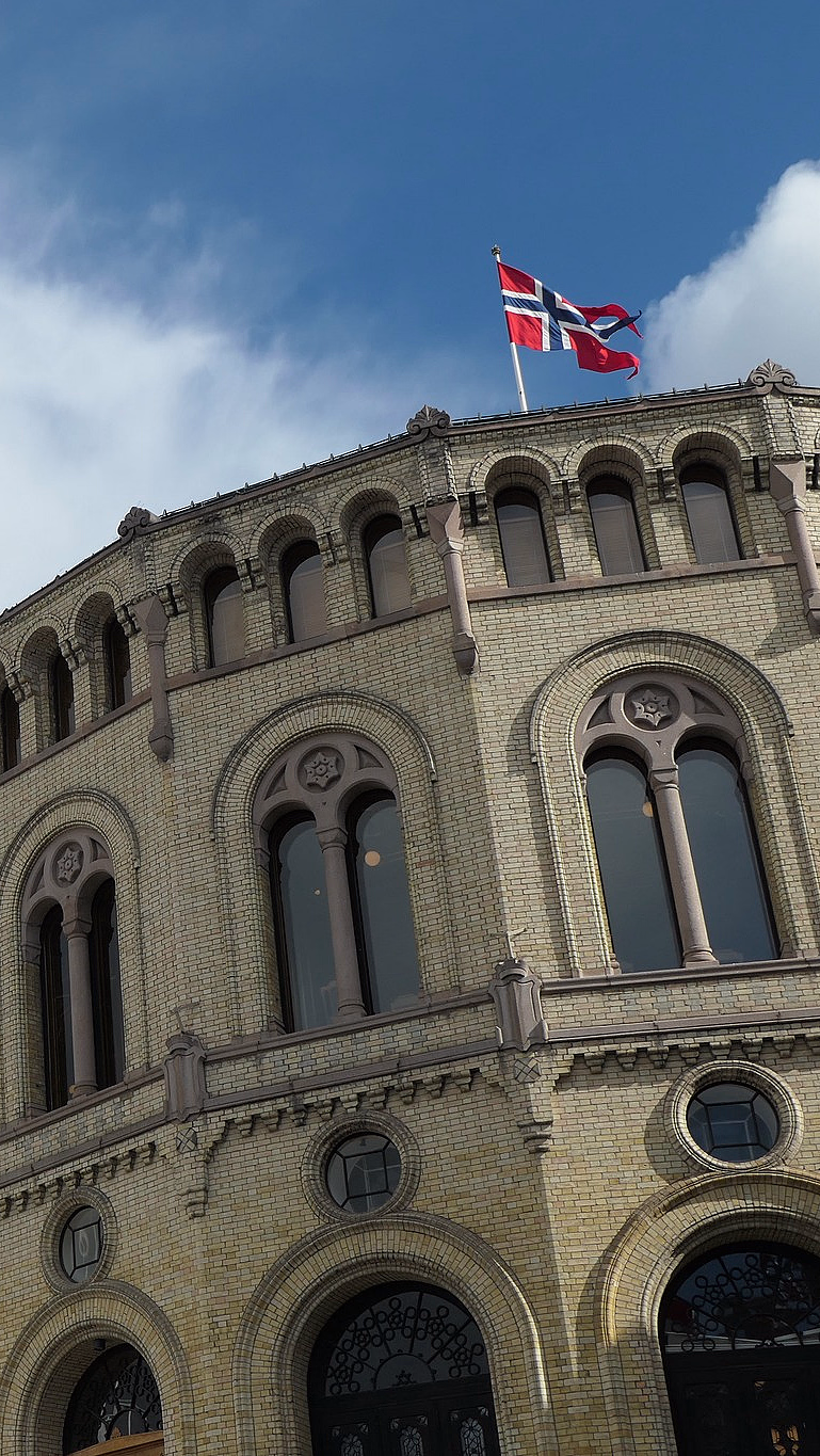 Stor løvestatue i stein foran en rund gul mursteinsbygning med buevinduer og det norske flagget på taket under en blå himmel.