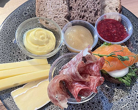 Grå spettet tallerken med brødskiver, ost, salami, røkelaks og små skåler med tilbehør på et trebord.