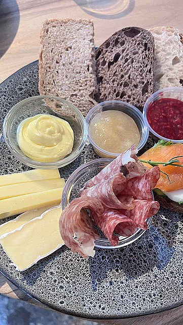 Grå spettet tallerken med brødskiver, ost, salami, røkelaks og små skåler med tilbehør på et trebord.