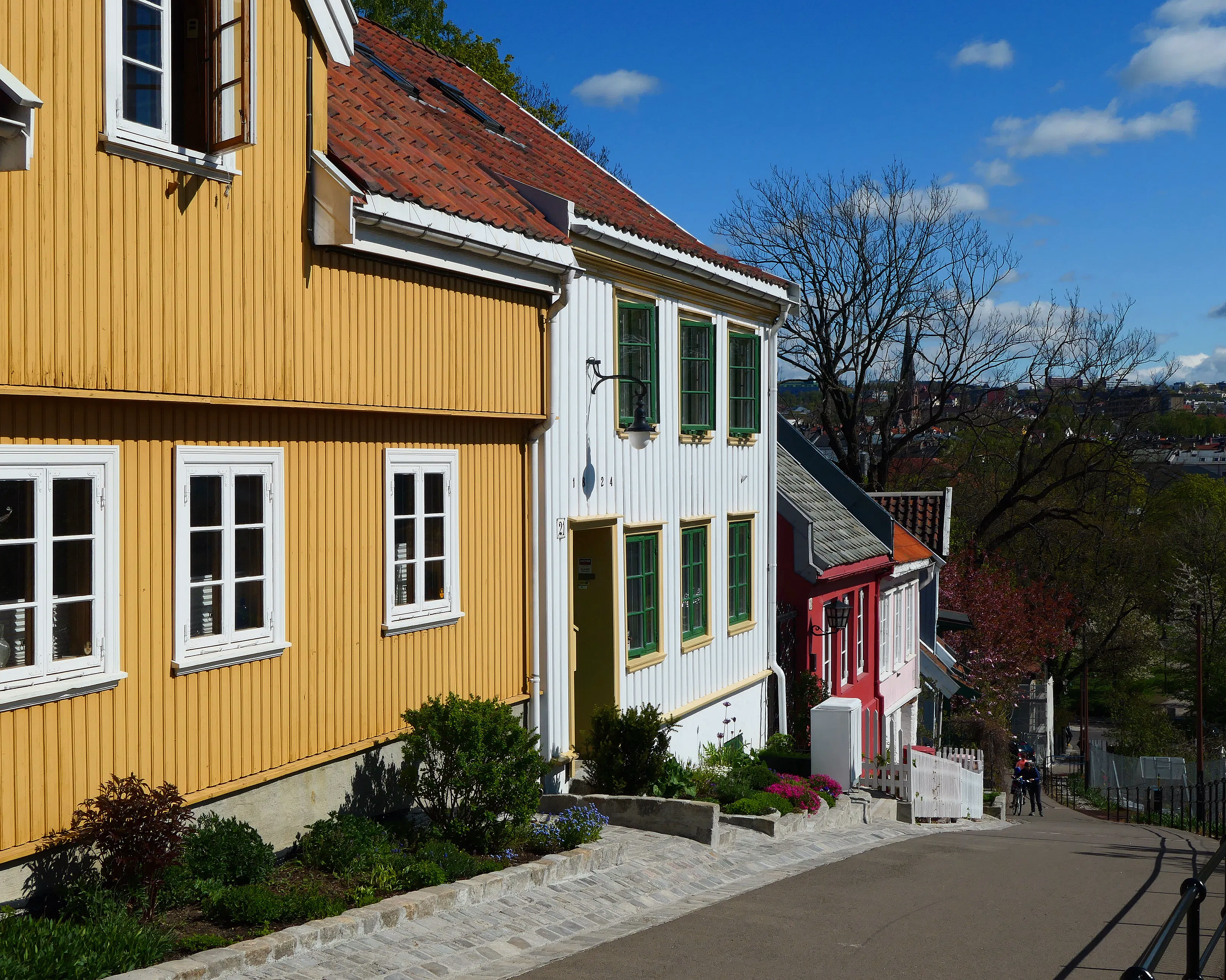 Bratt gate med fargerike trehus i gult, hvitt og rødt, blomsterbed langs veikanten og to syklister på vei ned bakken.
