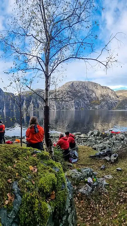 Gruppe med folk som hviler ved en fjord med høye fjell i bakgrunnen