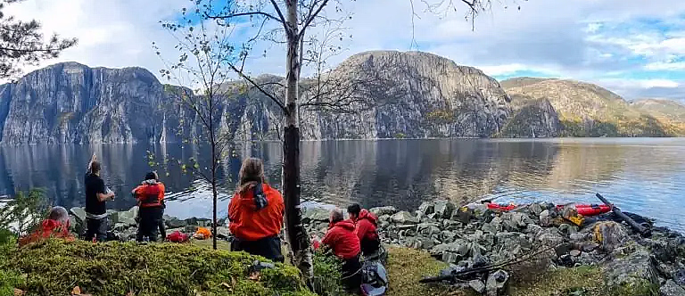 Gruppe med folk som hviler ved en fjord med høye fjell i bakgrunnen
