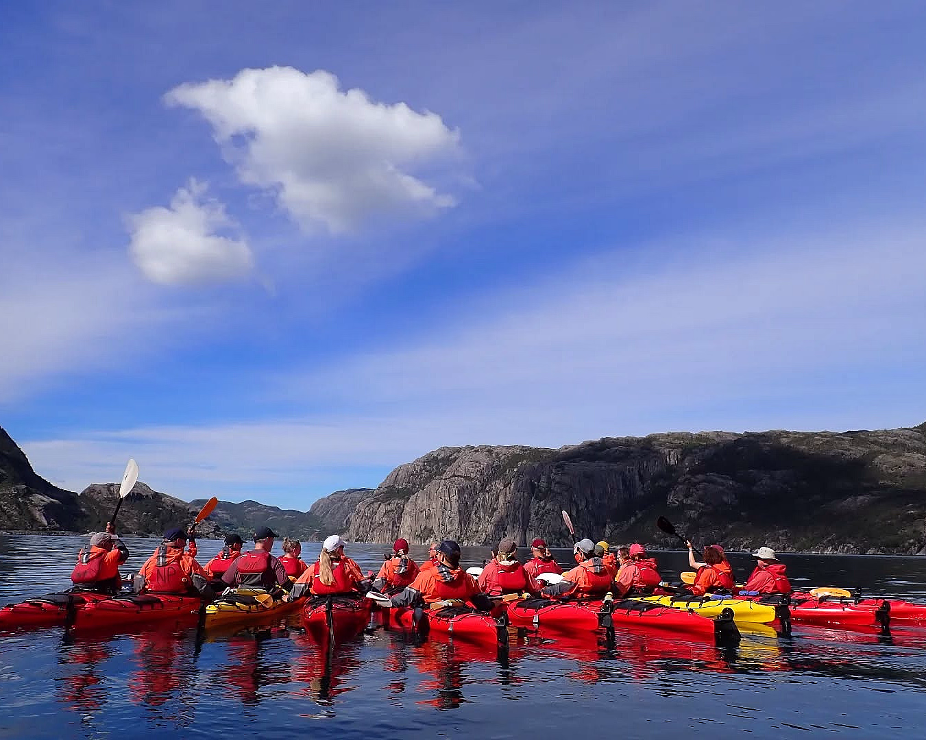 En stor gruppe padlere i røde jakker og fargerike kajakker på en rolig blå fjord, omgitt av bratte klipper og grønne fjellsider.