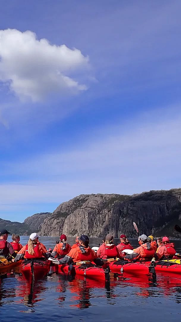 En stor gruppe padlere i røde jakker og fargerike kajakker på en rolig blå fjord, omgitt av bratte klipper og grønne fjellsider.