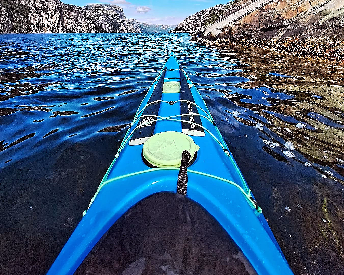 Blå kajakkbaug på mørkt vann i en fjord omgitt av bratte fjellvegger under en blå himmel med lette skyer.