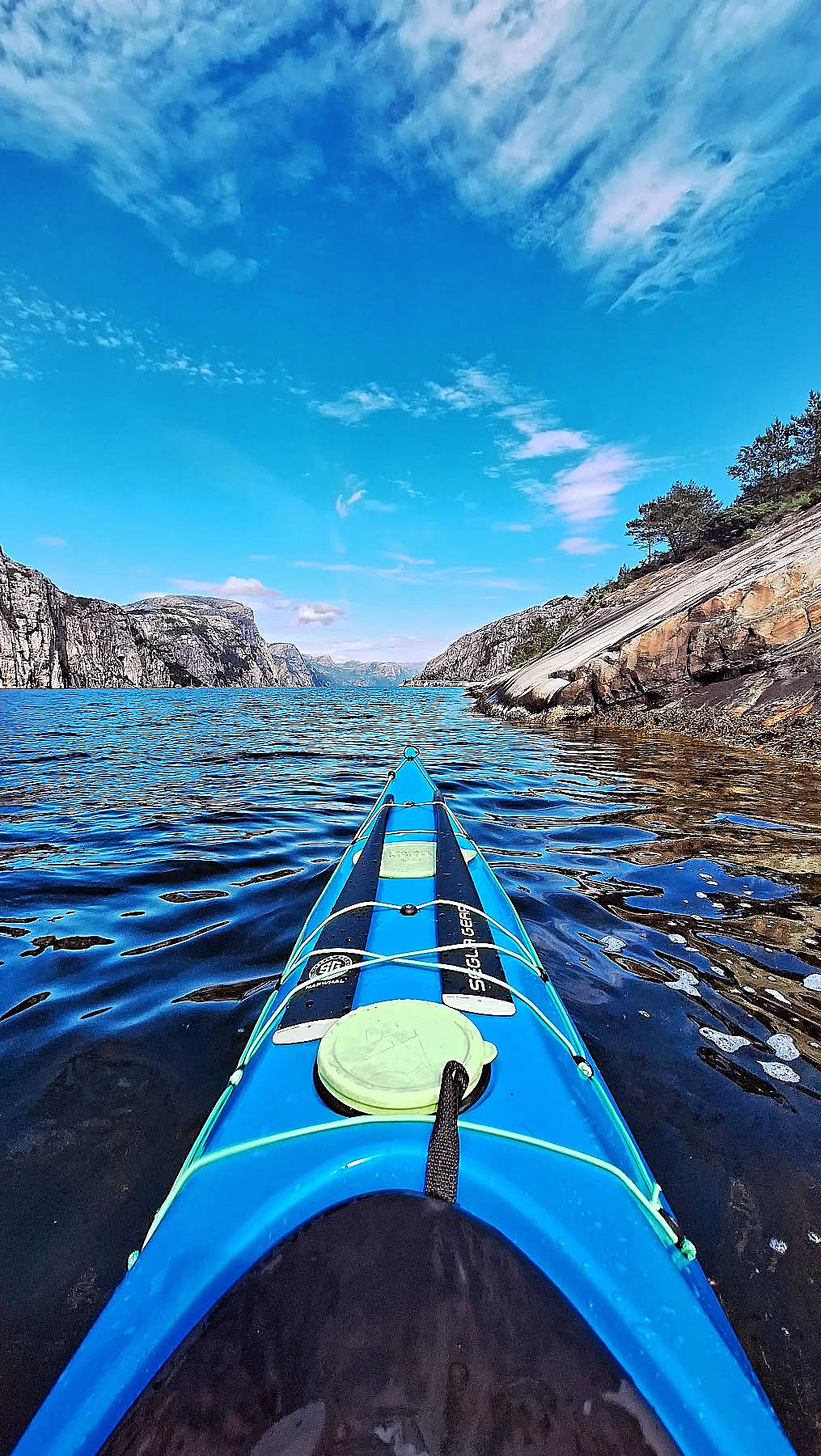 Blå kajakkbaug på mørkt vann i en fjord omgitt av bratte fjellvegger under en blå himmel med lette skyer.