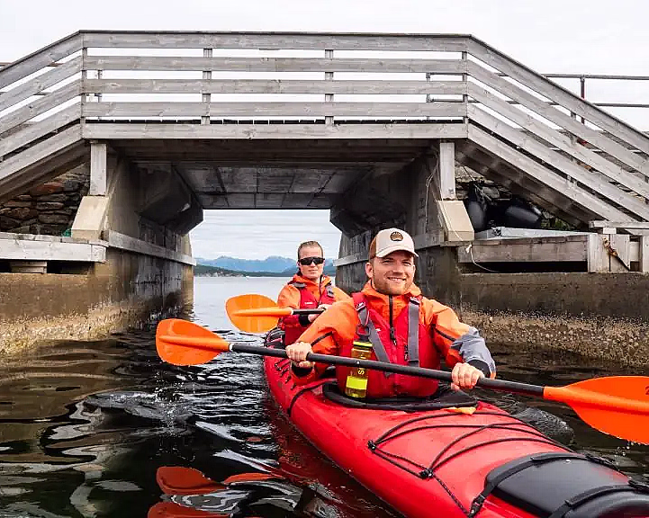 To personer i en rød tandemkajakk padler under en lav trebro i en smal kanal. De har på seg oransje redningsvester og bruker oransje årer, med blå åser og en overskyet himmel i bakgrunnen.