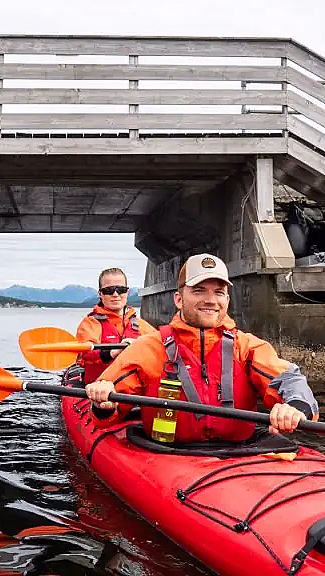 To personer i en rød tandemkajakk padler under en lav trebro i en smal kanal. De har på seg oransje redningsvester og bruker oransje årer, med blå åser og en overskyet himmel i bakgrunnen.