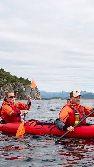 To personer i en rød tandemkajakk merket NORDIC PADDLING padler på rolig vann. De har på seg oransje redningsvester og bruker oransje årer, med fjell og grønn kyst i bakgrunnen.