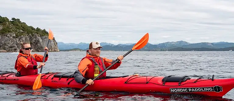 To personer i en rød tandemkajakk merket NORDIC PADDLING padler på rolig vann. De har på seg oransje redningsvester og bruker oransje årer, med fjell og grønn kyst i bakgrunnen.