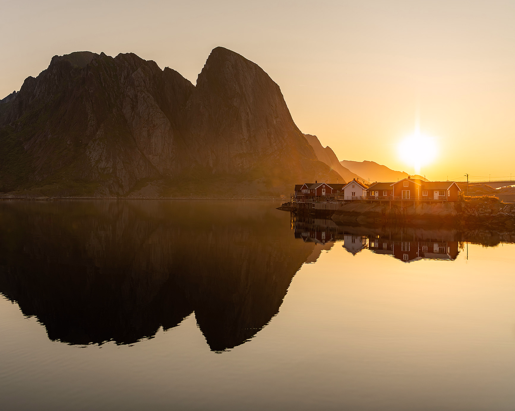 En rød ørken ved soloppgang med to store, flate fjellformasjoner i bakgrunnen. I forgrunnen er en tørr busk på sprukken jord med små steiner og lange blå skygger under en klar himmel.