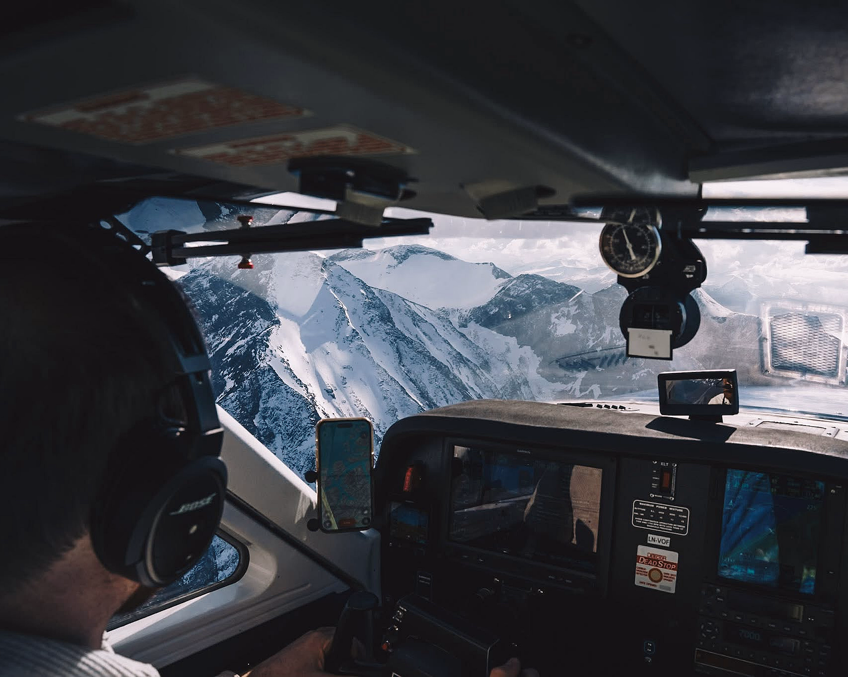 Innsiden av en flycockpit med instrumentpanel og en pilot ved spakene, med utsikt over snødekte fjell og en blå innsjø gjennom frontruten.