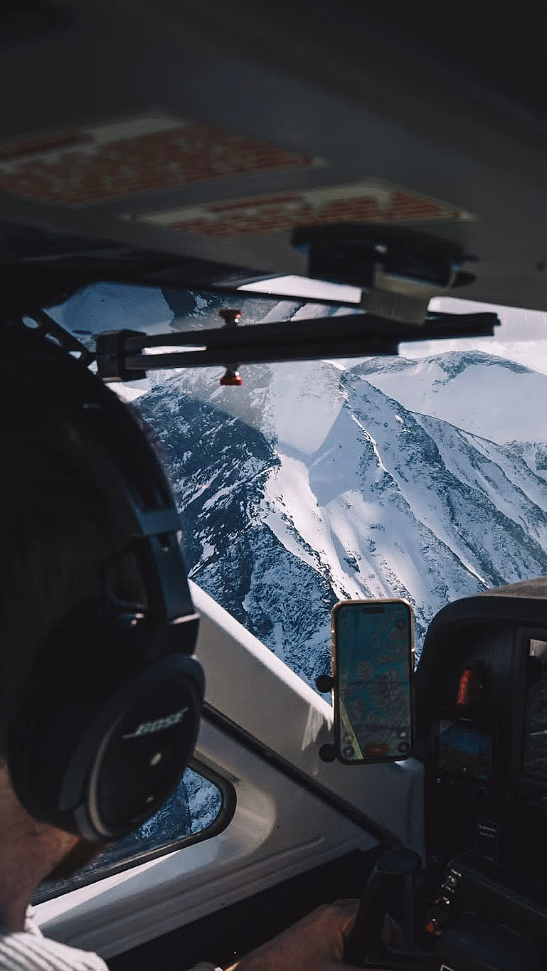 Innsiden av en flycockpit med instrumentpanel og en pilot ved spakene, med utsikt over snødekte fjell og en blå innsjø gjennom frontruten.
