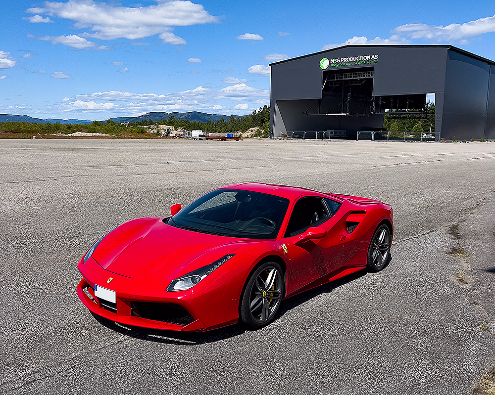 Skinnende rød Ferrari sportsbil parkert på grå asfalt foran en stor hangar under en blå himmel.