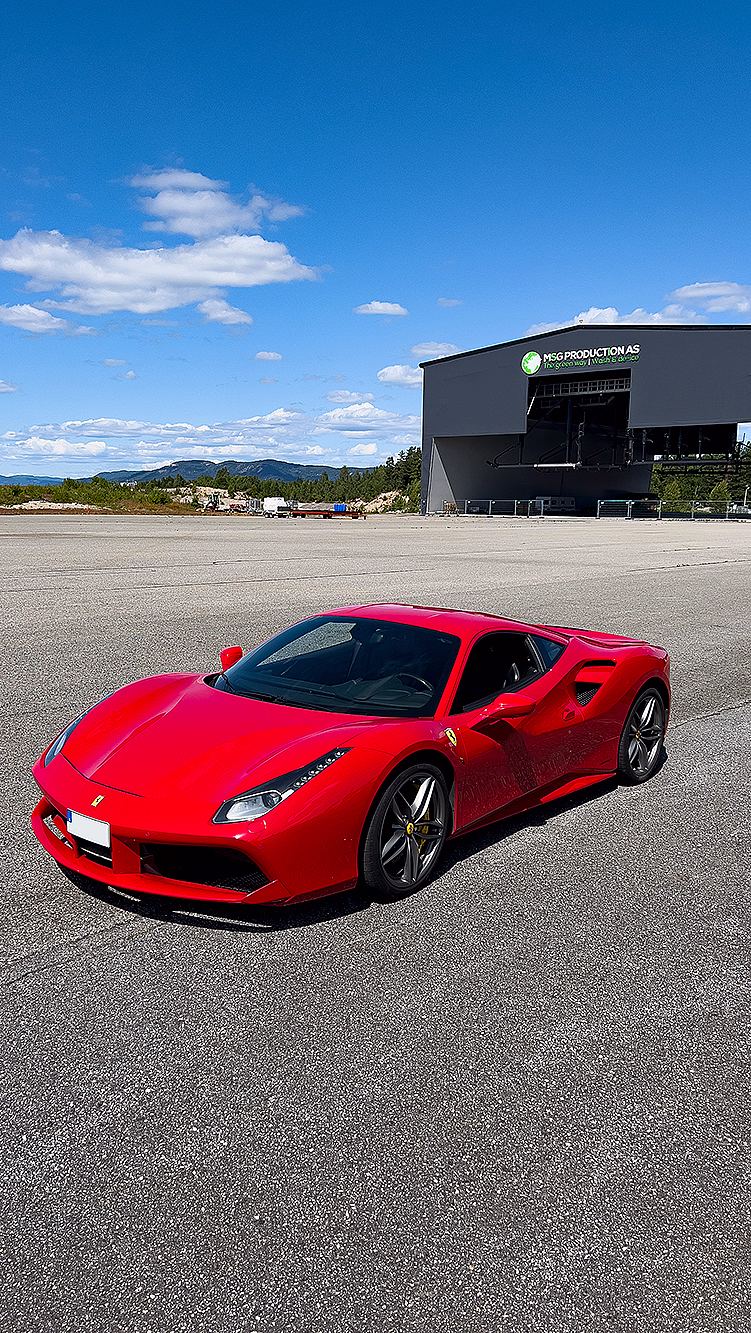 Skinnende rød Ferrari sportsbil parkert på grå asfalt foran en stor hangar under en blå himmel.