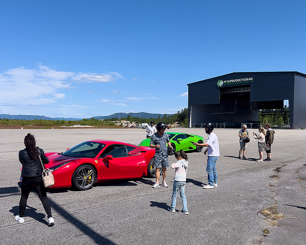En rød Ferrari og en grønn sportsbil parkert på en asfaltert plass foran en stor grå bygning på en solfylt dag.