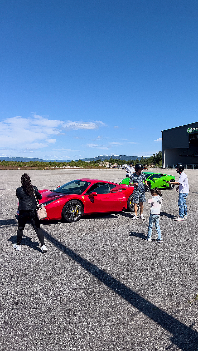 En rød Ferrari og en grønn sportsbil parkert på en asfaltert plass foran en stor grå bygning på en solfylt dag.
