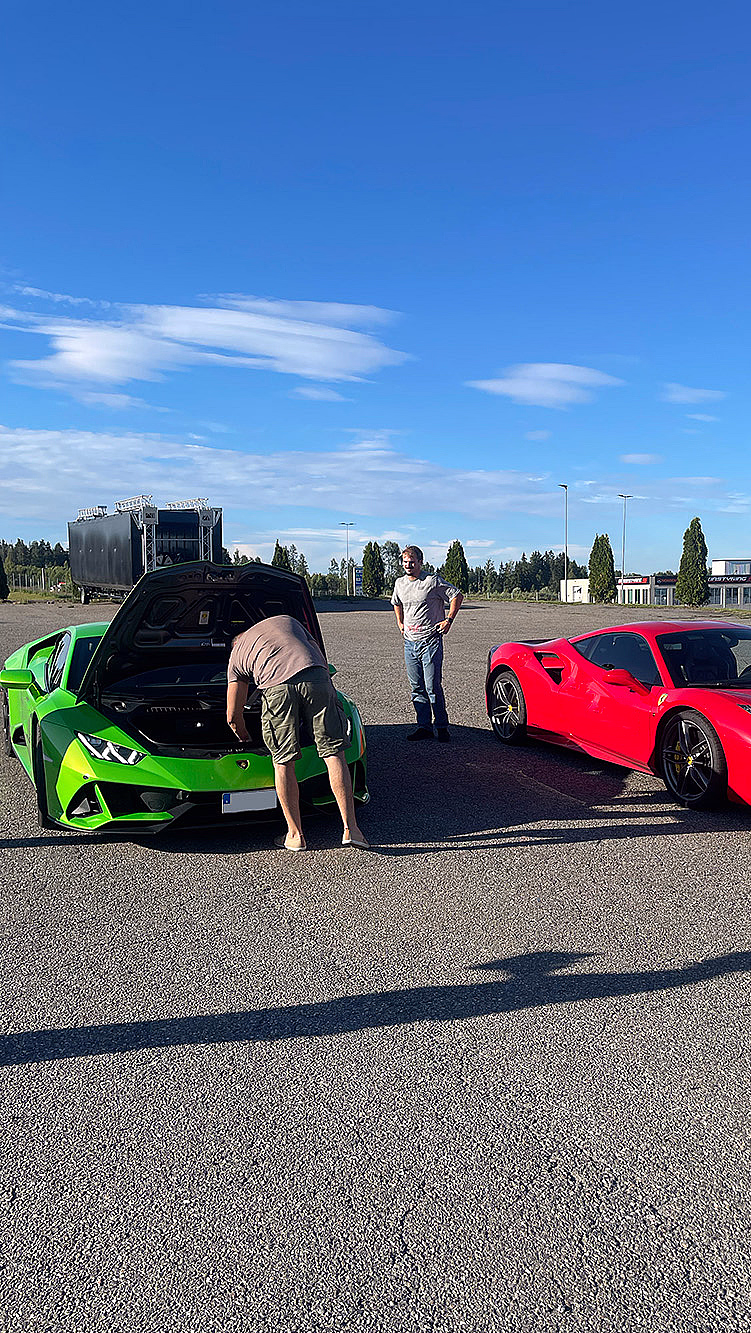 En lysegrønn Lamborghini med åpent bagasjerom foran og en rød Ferrari parkert på en asfaltert plass under en blå himmel.