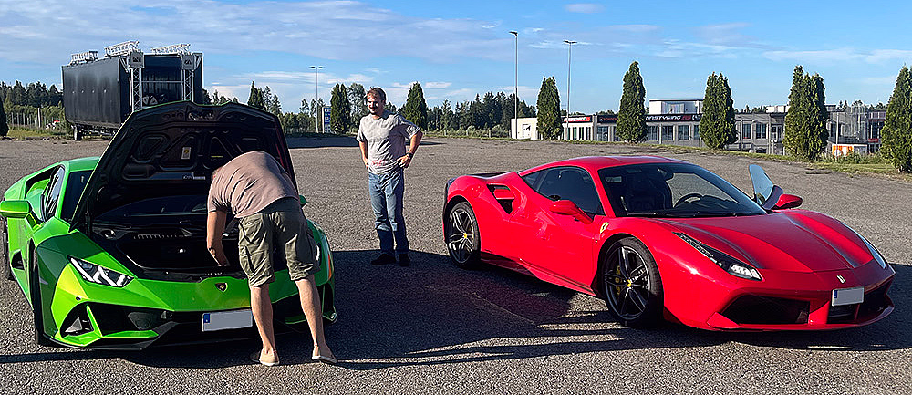 En lysegrønn Lamborghini med åpent bagasjerom foran og en rød Ferrari parkert på en asfaltert plass under en blå himmel.