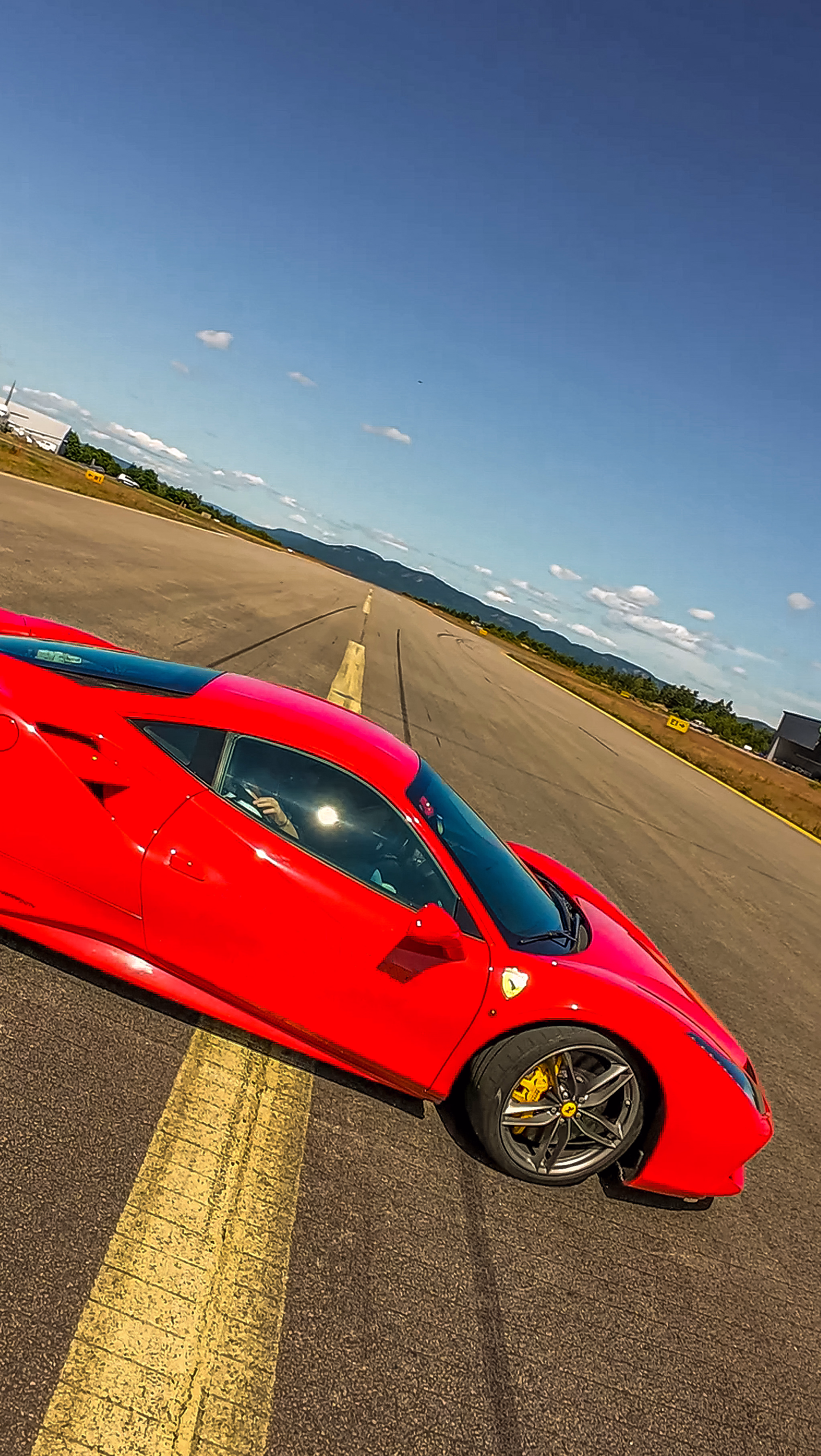 Sideprofil av en rød Ferrari sportsbil på en rullebane med en gul midtlinje, grå felger og gule bremsekalipere under en blå himmel.