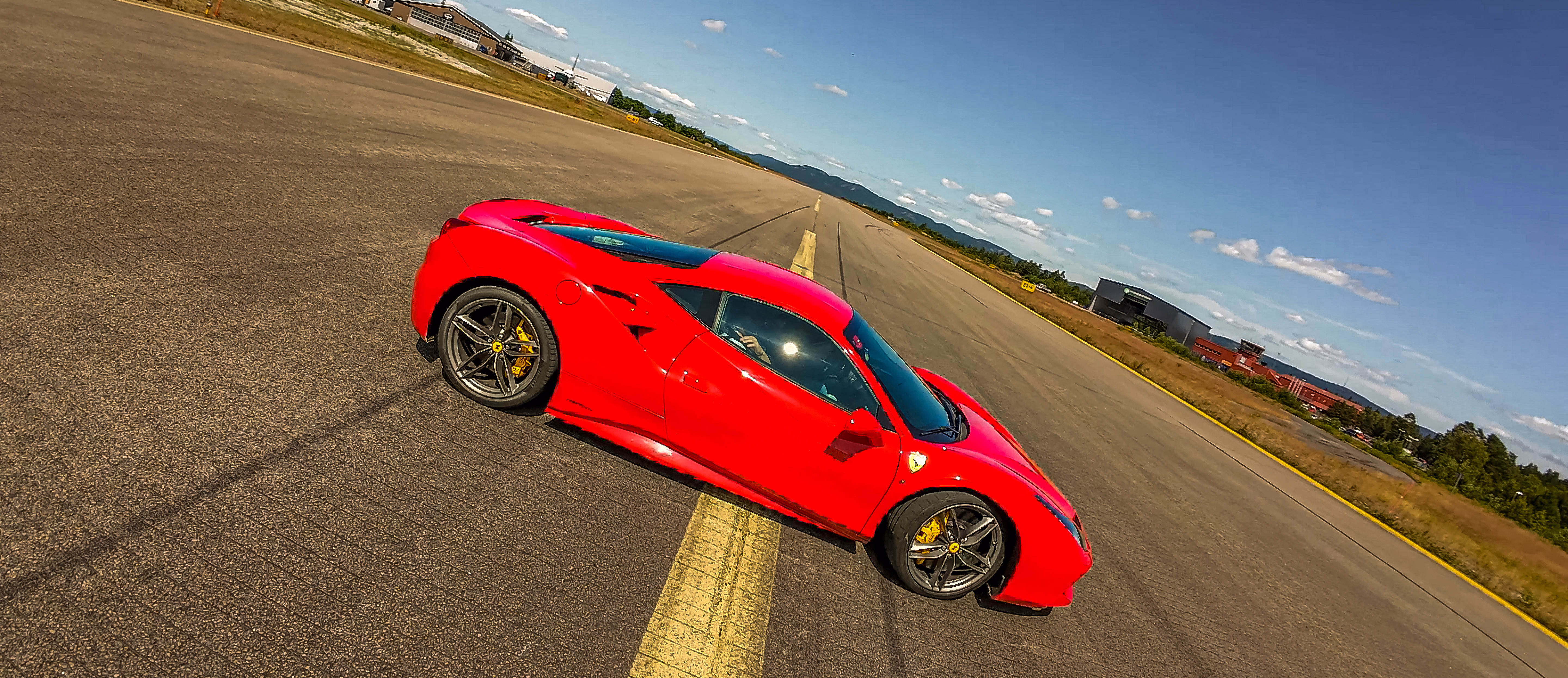 Sideprofil av en rød Ferrari sportsbil på en rullebane med en gul midtlinje, grå felger og gule bremsekalipere under en blå himmel.
