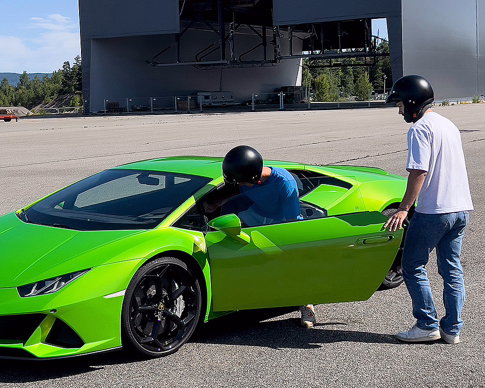 En limegrønn sportsbil med svarte felger på en asfaltert plass. Førerdøren er åpen, og to personer med hjelmer står ved bilen foran en stor grå hangar med skiltet "MSG PRODUCTION AS".