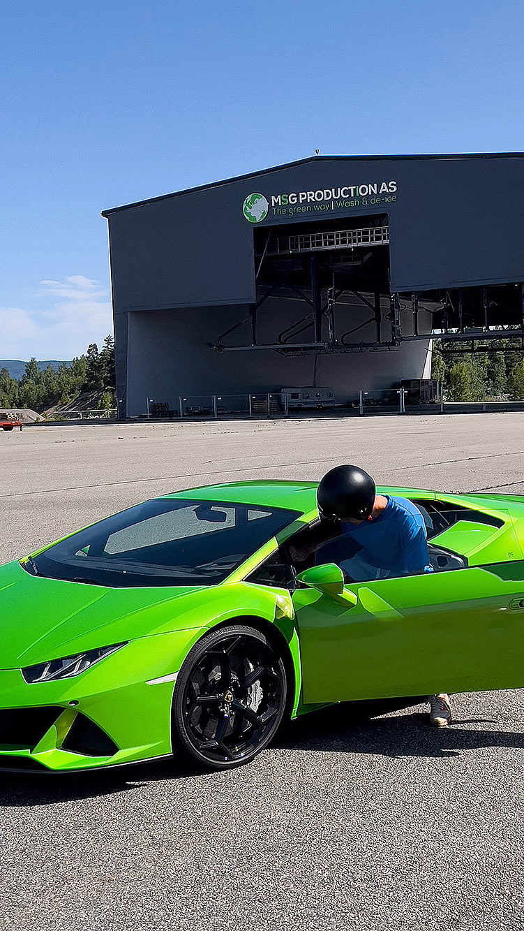 En limegrønn sportsbil med svarte felger på en asfaltert plass. Førerdøren er åpen, og to personer med hjelmer står ved bilen foran en stor grå hangar med skiltet "MSG PRODUCTION AS".
