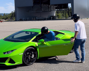 En limegrønn sportsbil med svarte felger på en asfaltert plass. Førerdøren er åpen, og to personer med hjelmer står ved bilen foran en stor grå hangar med skiltet "MSG PRODUCTION AS".