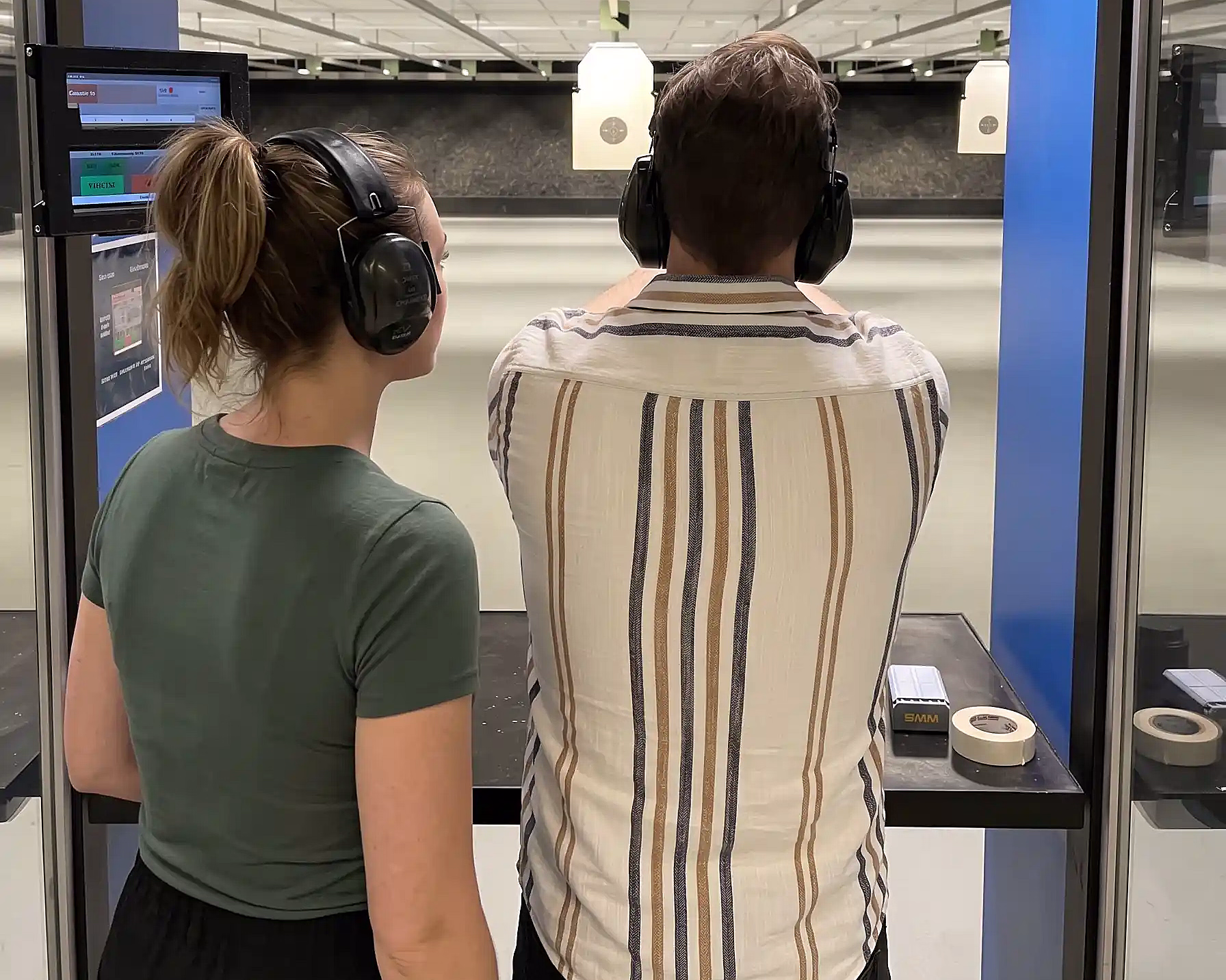 To personer sett bakfra på en innendørs skytebane, begge med svarte hørselvern. Den ene sikter mot hengende blinker, og på en svart benk i forgrunnen ligger en ammunisjonseske og taperuller.