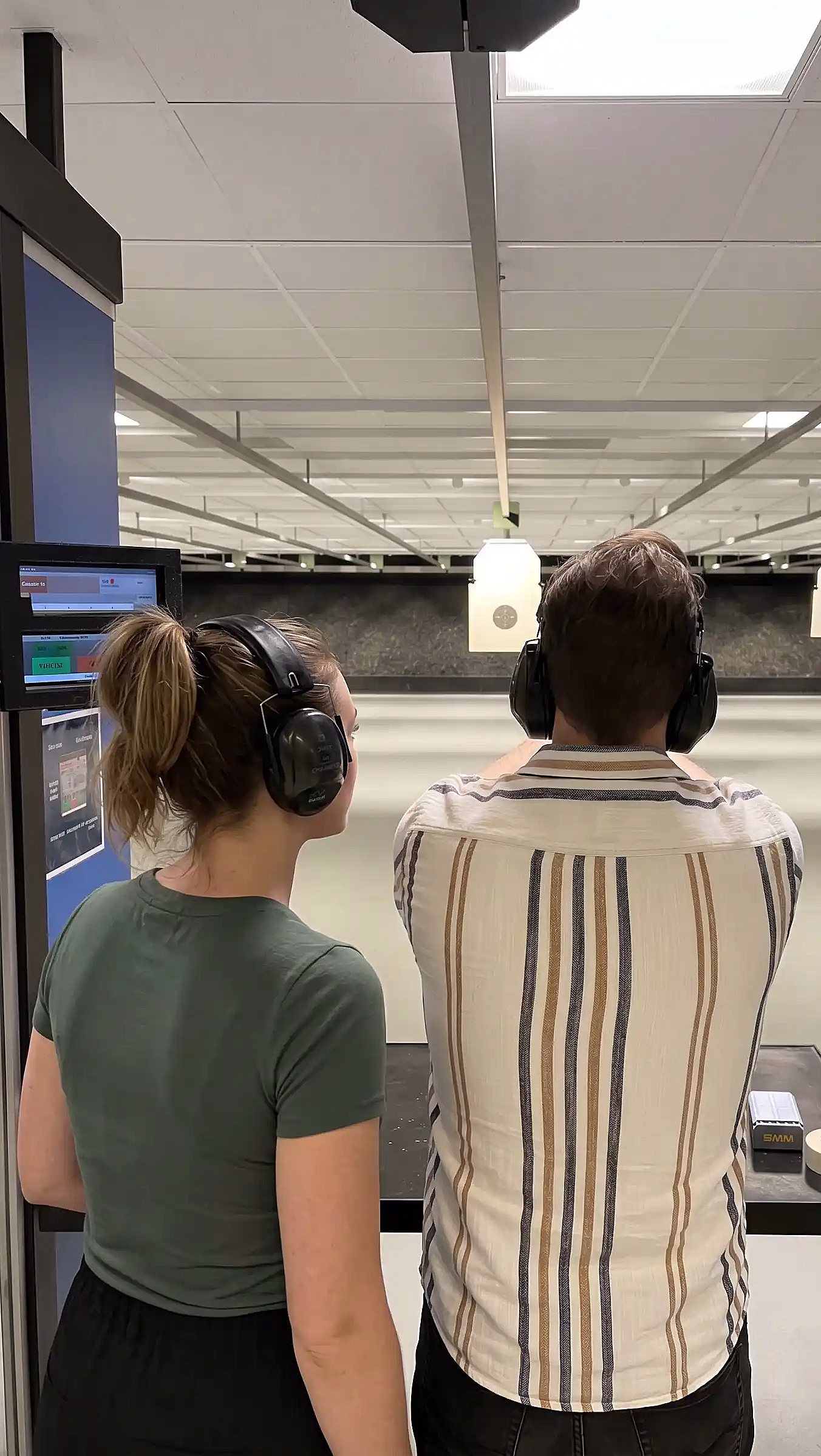 To personer sett bakfra på en innendørs skytebane, begge med svarte hørselvern. Den ene sikter mot hengende blinker, og på en svart benk i forgrunnen ligger en ammunisjonseske og taperuller.