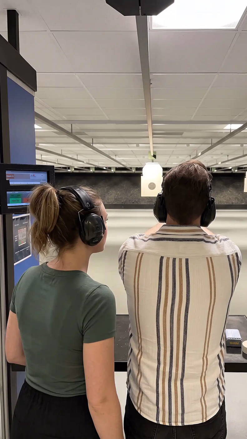 To personer sett bakfra på en innendørs skytebane, begge med svarte hørselvern. Den ene sikter mot hengende blinker, og på en svart benk i forgrunnen ligger en ammunisjonseske og taperuller.