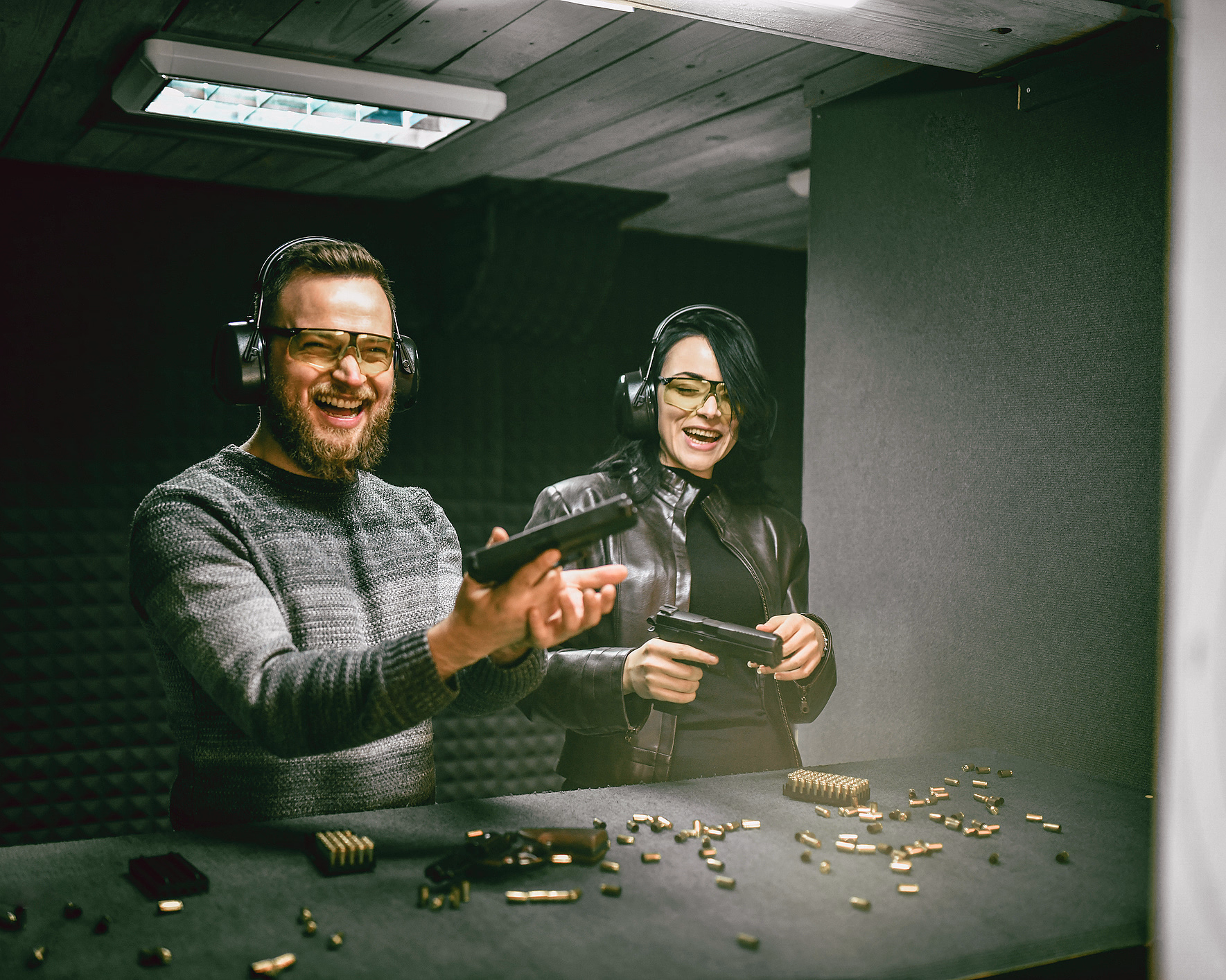 To personer med hørselvern og gule vernebriller holder pistoler på en innendørs skytebane.