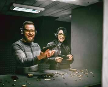To personer med hørselvern og gule vernebriller holder pistoler på en innendørs skytebane.