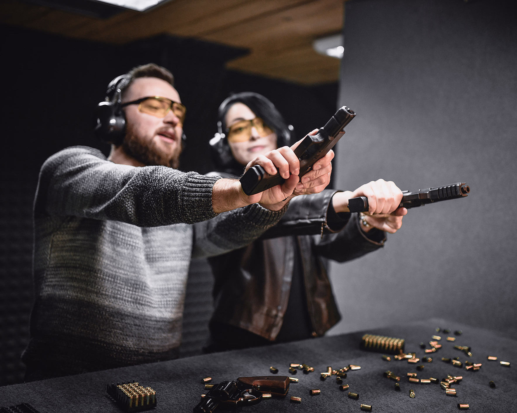 To voksne på en innendørs skytebane med hørselvern og gule vernebriller, som sikter med pistoler over et bord fylt med ammunisjon og en revolver.
