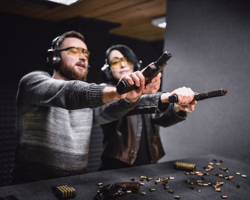 To voksne på en innendørs skytebane med hørselvern og gule vernebriller, som sikter med pistoler over et bord fylt med ammunisjon og en revolver.