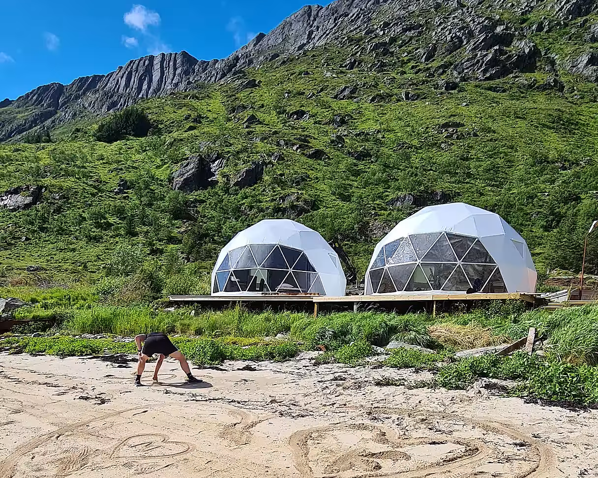 To hvite glampingdomer på en treplatting ved en strand med fjell i bakgrunnen