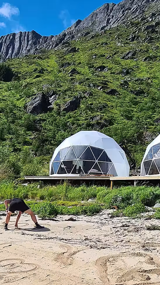 To hvite glampingdomer på en treplatting ved en strand med fjell i bakgrunnen