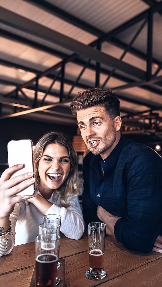 Mann og kvinne tar en selfie ved et bord med ølglass i et lokale med industritak
