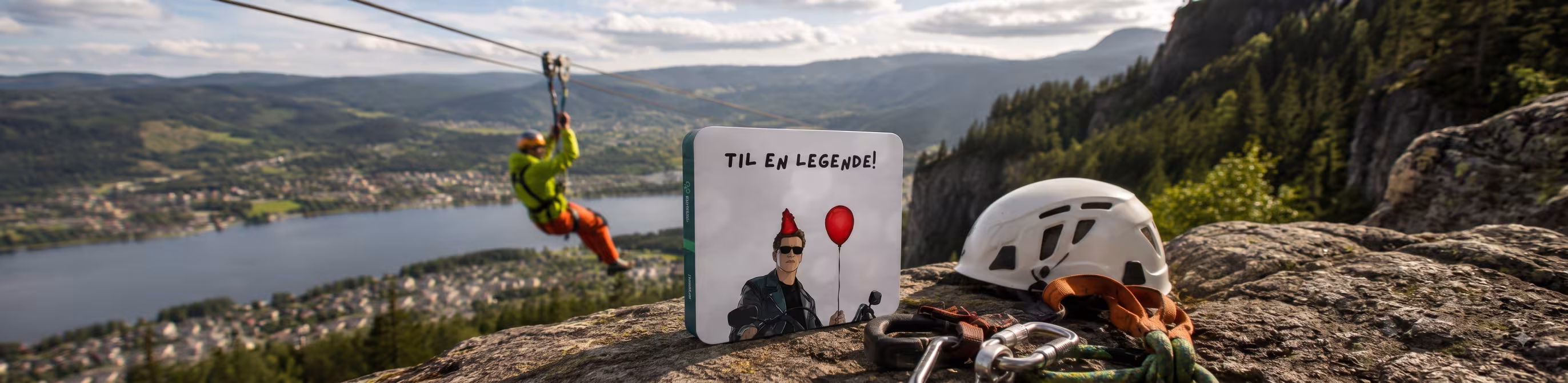 Person i zipline over en fjord med en gaveeske og klatreutstyr i forgrunnen