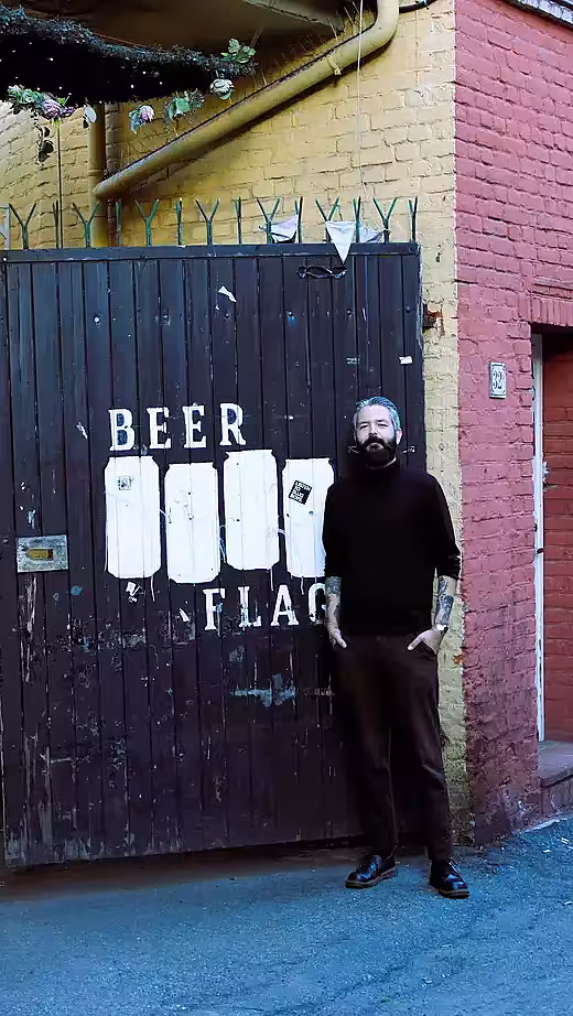 En mann med skjegg står foran en mørk treport med teksten Beer Flag