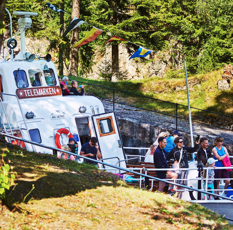 Hvit båt med navnet Telemarken i en kanal med folk på dekk og fargerike flagg