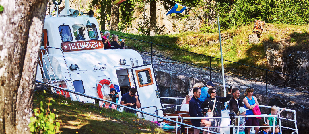 Hvit båt med navnet Telemarken i en kanal med folk på dekk og fargerike flagg