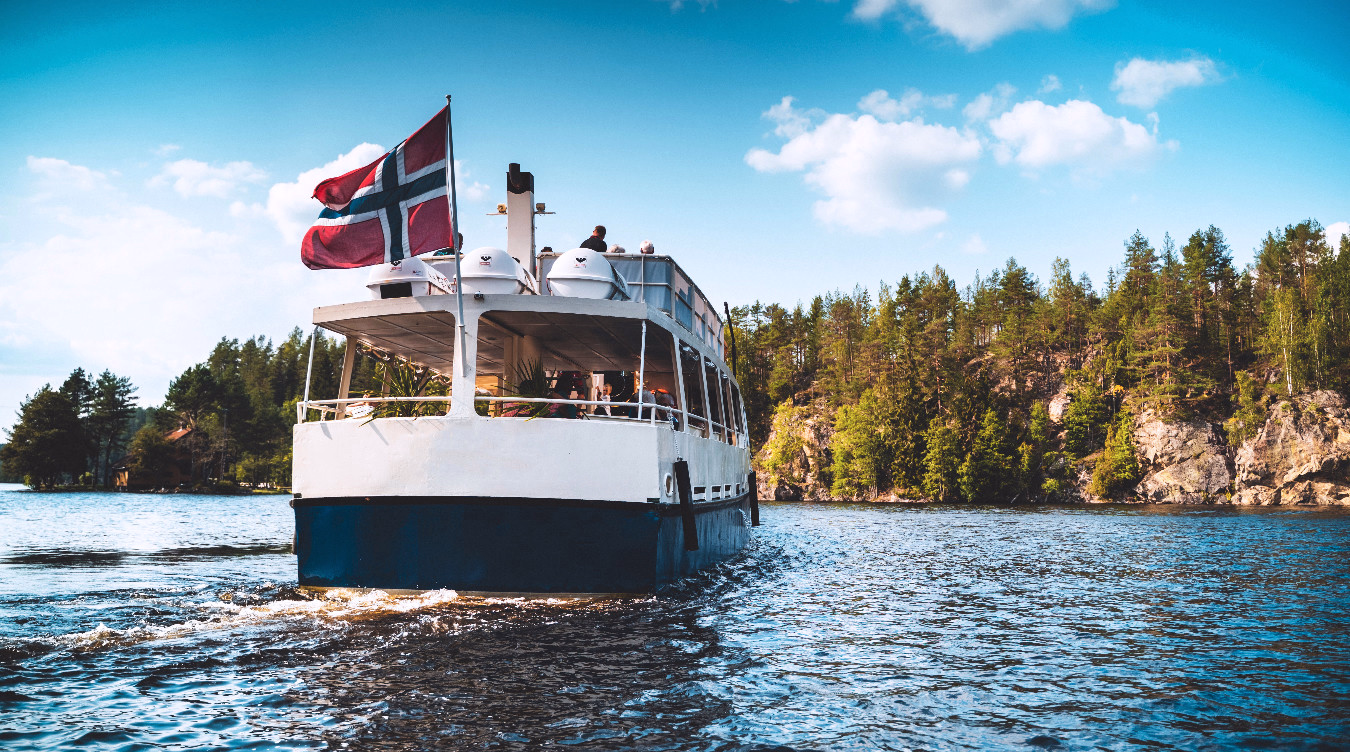 Hvit og blå båt med passasjerer på en innsjø med skog og klipper i bakgrunnen