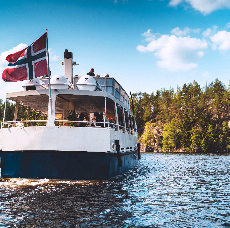 Hvit og blå båt med passasjerer på en innsjø med skog og klipper i bakgrunnen
