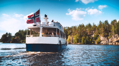 Hvit og blå båt med passasjerer på en innsjø med skog og klipper i bakgrunnen